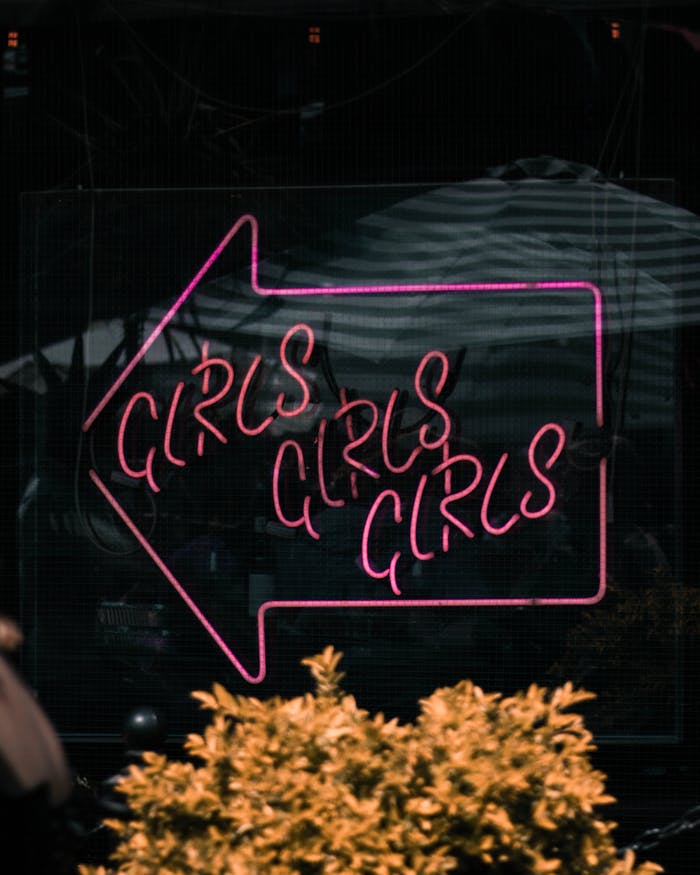 Pink neon 'Girls Girls Girls' sign in an outdoor street setting.